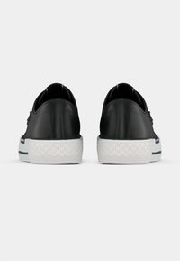 Τα μαύρα αθλητικά παπούτσια διαθέτουν λείο υλικό, με χοντρή λευκή σόλα και διακριτικές μαύρες και μπλε λεπτομέρειες. Το πίσω μέρος παρουσιάζει καθαρό ράψιμο και υφή στη φτέρνα.