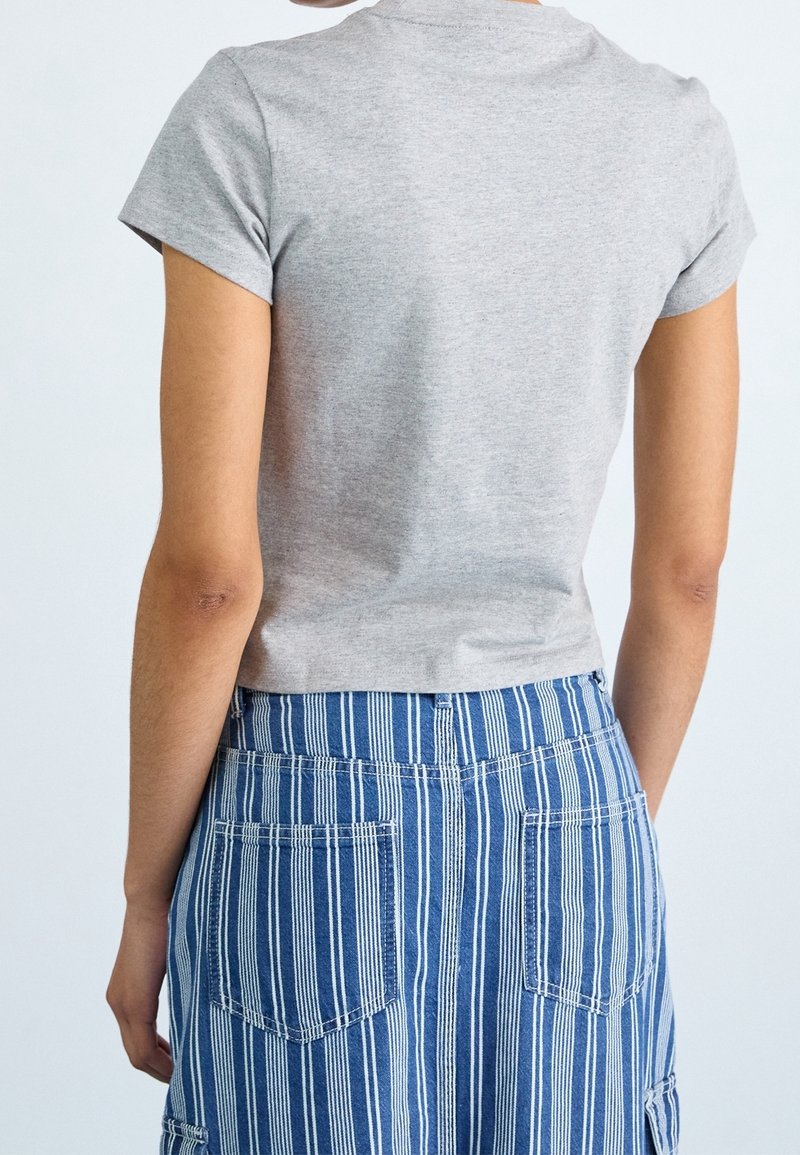 Persona con una camisa de manga corta gris claro y pantalones de rayas verticales azules y blancas, vista desde atrás contra un fondo liso.