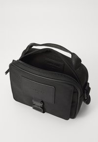 Black fabric crossbody bag with a zipper closure, top handle, and front patch. Textured surface with a smooth leather accent.