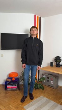 Fekete zipzáras kapucnis pulóver sárga logóval, kék farmernadrág és sötétbarna cipő, élénk színű belső térben dekoratív kiegészítők mellett.