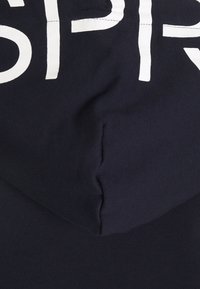 Tela de sudadera con capucha de color azul marino con una textura suave; presenta grandes letras blancas en la parte frontal y una capucha redondeada.