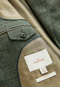 Tamsiai žalia tekstūruota audinio švarkas su smėlinės spalvos pamušalu, mėlynos spalvos akcento juostele, sagų detalėmis ir matomais priežiūros etiketėmis vidinėje pusėje.