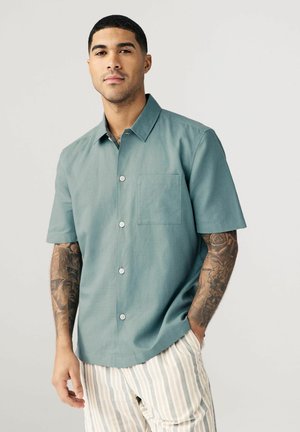 Short-sleeve shirt in muted teal, lightweight fabric, button-front, with a chest pocket. Paired with striped pants in soft tones.