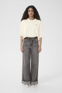 Sieviete ar cirtainiem matiem, valkājot pārāk lielu krēmkrāsas polo krekli, augstās vidukļa pelēkās plataji džinsa bikses ar jostu un baltas smailas kurpes, stāvot.