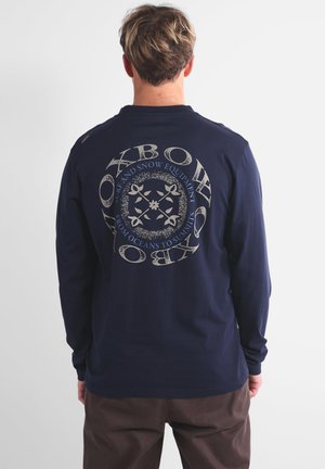 Chemise à manches longues navy en coton avec un graphique circulaire au dos, présentant du texte et des motifs intriqués en beige clair.