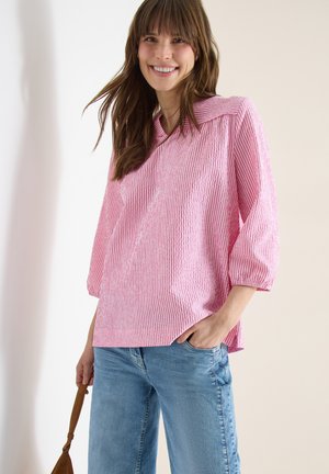 Smilende kvinde iført en pink og hvidstribet bluse med 3/4-ærmer og lyseblå jeans, der holder i håndtaget på en brun taske.