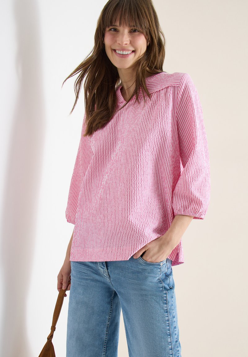 Mujer sonriente con una blusa de rayas rosas y blancas de manga tres cuartos y jeans azul claro, sosteniendo el asa de una bolsa marrón.