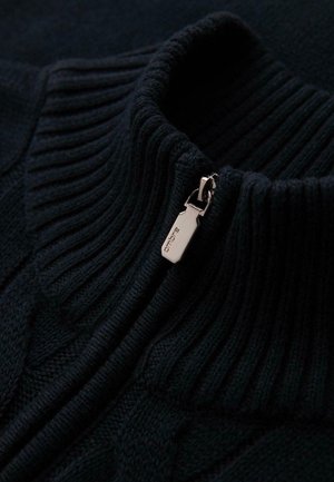 Gros plan sur le col d'un pull en maille ribbed noir avec un tirette de fermeture éclair en argent gravée du nom de la marque "ombre."