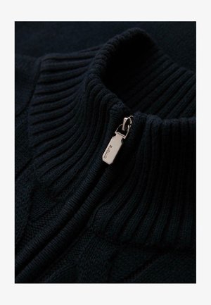 Gros plan sur le col d'un pull en maille ribbed noir avec un tirette de fermeture éclair en argent gravée du nom de la marque "ombre."