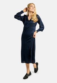 Rochie midi bleumarin din material mătăsos cu model texturat, având mâneci lungi și decolteu în V. Purtați cu mocasini negri.