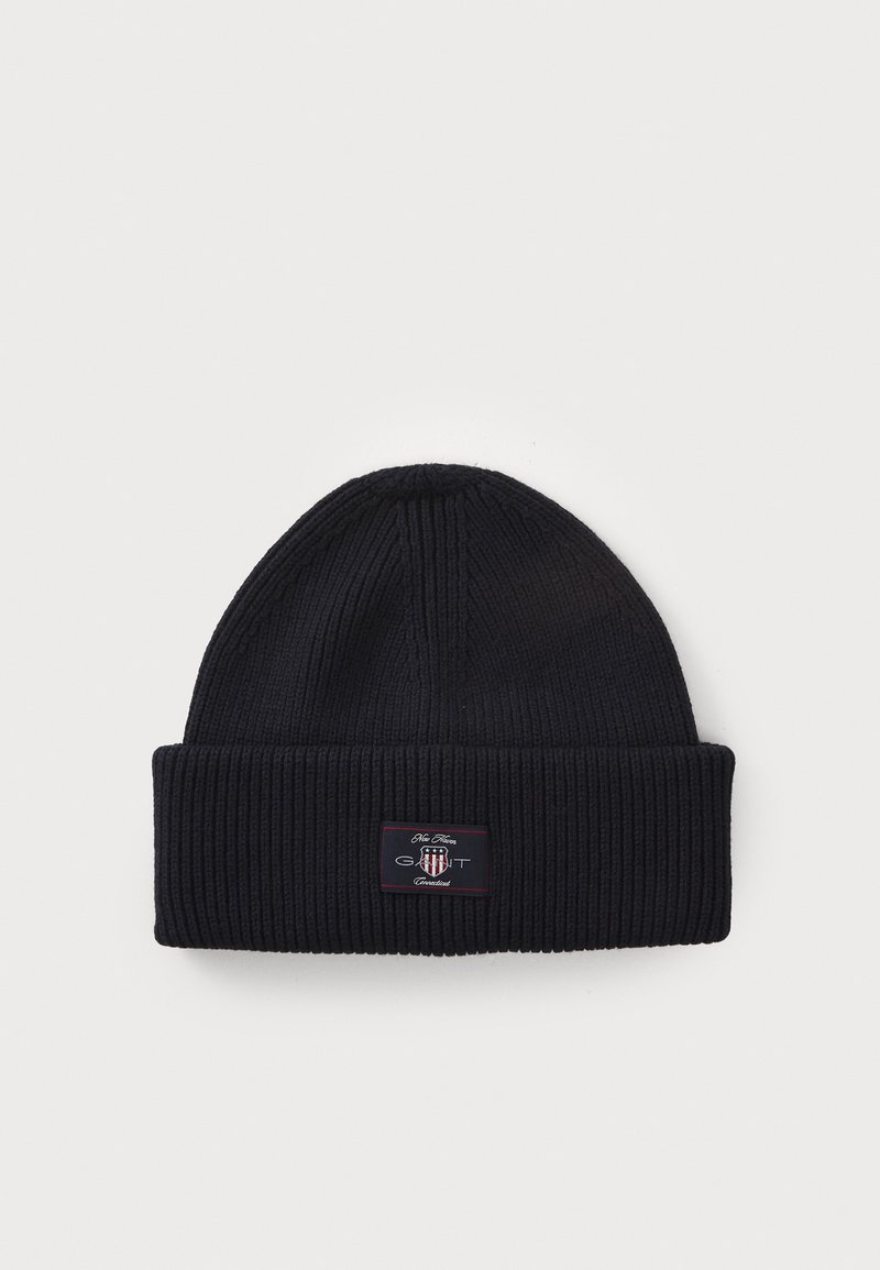 Námornícky modrá pletená čiapka, rebrovaná textúra, prehnutý lem, s malou značkovou nášivkou v červenej a bielej farbe na prednej strane.