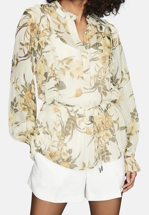 Vrouw draagt een doorschijnende, lange mouwen bloemenblouse met ruches en witte shorts, staand met ontspannen handen langs het lichaam.