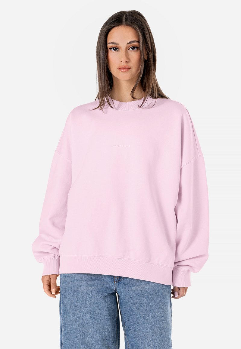 Felpa oversize rosa realizzata in morbido tessuto, con scollo tondo e polsini a costine, abbinata a jeans in denim azzurri.