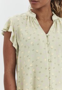 Grüne Bluse mit floralem Muster, kurzen Rüschensäumen, Knopfleiste und gerafftem Ausschnitt. Strukturstoff mit lila und gelben Akzenten.