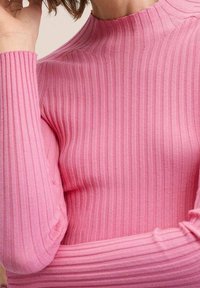 Dopasowana bluzka z długim rękawem o prążkowanej fakturze, z wysokim kołnierzem. Materiał jest miękki, w jednolitym jasnoróżowym kolorze.