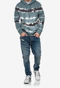 Hombre con sudadera con capucha teñida anudada en tonos teal, negro y blanco con bolsillo frontal, vaqueros azules desgastados y zapatillas blancas con detalles negros, con las manos en los bolsillos.