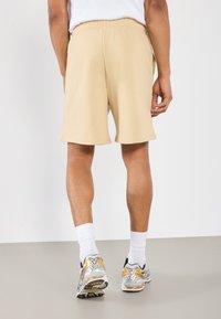Shorts de sport beiges avec une taille élastique, texture lisse et longueur mi-cuisse, associés à des chaussettes blanches et des baskets multicolores.