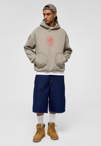 Beige hoodie met cirkelvormige rode graphic, met grote zijzakken. Draagt het met donkerdenim shorts en gele werklaarzen.