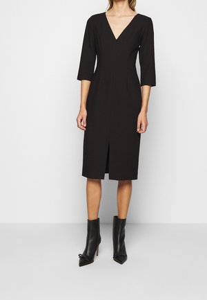 Kobieta ubrana w czarną sukienkę do kolan z rękawami o długości do łokcia i dekoltem w kształcie litery V, połączoną z czarnymi botkami na obcasie z zaokrąglonym noskiem.