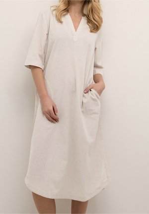 Kvinde iført en knælængde, løstsiddende beige kjole med albuelange ærmer, V-hals og sidelommer, stående mod en ensfarvet baggrund.