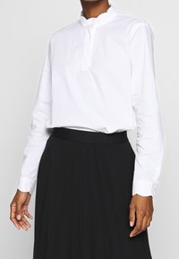 Vit långärmad blus med hög krage, med en partiell knapplist och slät textur, bärs tillsammans med en svart plisserad kjol.