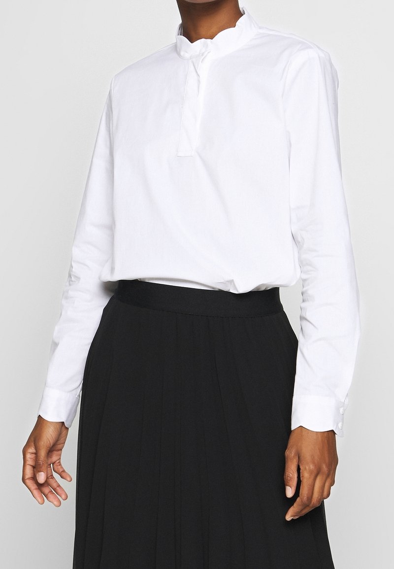 Vit långärmad blus med hög krage, med en partiell knapplist och slät textur, bärs tillsammans med en svart plisserad kjol.