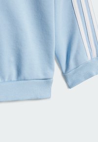 Felpa azzurro chiaro in tessuto, con polsini a costine, caratterizzata da tre strisce bianche lungo un lato. Texture morbida e design informale.