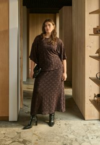 Femme en robe marron texturée et bottes noires, debout dans un couloir aux murs en bois clair et au sol en marbre, tenant un sac à main noir.