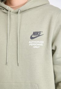 Olivno zelena Nike kapuca z vrvicami. Ponuja napis "DOVOLJENI SAMO POOBLAŠČENI" v beli barvi. Mehka tkanina z udobnim krojem in sprednjim žepom.