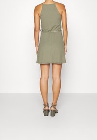 Olivově zelené šaty bez rukávů s vypasovaným pasem, rozšířenou sukní a texturovanou tkaninou. Nosené s olivově zelenými sandály na blokovém podpatku.