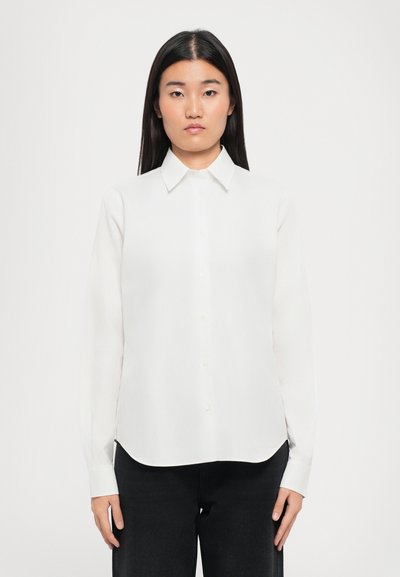 Chemise blanche à manches longues, avec col, léger éclat et ourlet arrondi, associée à un pantalon sombre. Tissu uni, sans motifs.