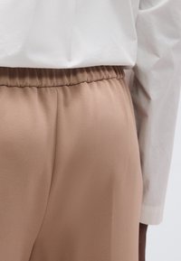 Μπεζ κωνικό παντελόνι από υφασμάτινη υφή, με ελαστική μέση. Ένα λευκό πουκάμισο με μακριά μανίκια φαίνεται μερικώς από πάνω.