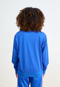 Persoon met krullend haar in een blauw trainingspak met roze strepen staat met de rug naar een effen witte achtergrond.