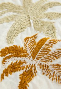 Broderie de palmier beige et orange sur tissu blanc texturé, montrant des motifs de fils détaillés et des coutures en relief.