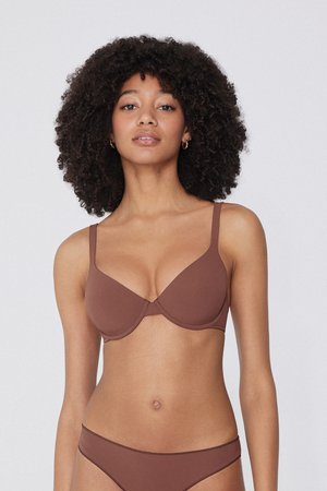 Mujer con cabello rizado, vestida con un sujetador marrón y ropa interior a juego, de pie frente a un fondo claro y sencillo.
