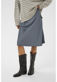 Jupe en satin gris avec une texture lisse, associée à des bottes de cowboy en cuir noir. La tenue est complétée par un pull rayé beige et gris.
