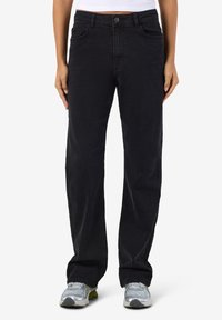 Oseba, oblečena v črne naravnost krojene hlače z visokim pasom in sprednjimi žepi ter srebrno-rumenimi supergami, stoji ob belem ozadju.