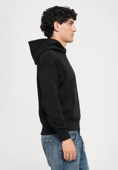 Uomo con felpa nera e jeans blu in piedi di profilo contro uno sfondo bianco semplice.