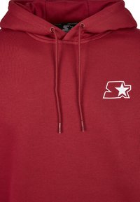 Rød hettegenser laget av mykt stoff, med frontlomme, snorhette og en hvit brodert logo med en stjerne.
