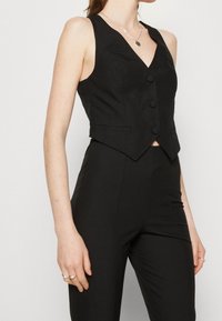 Svart ärmlös västtopp med v-ringning, som har tre knappar och en kortad design. Slät tyg och åtsittande silhuett.