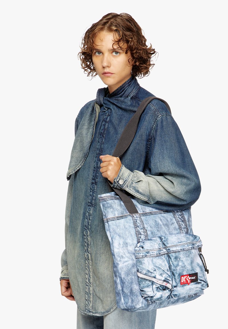 Denim-kangaskassi vaaleansinisellä värillä, jossa on edessä vetoketjuluukku, harmaa hihna ja säädettävä muotoilu. Sisältää teksturoituja yksityiskohtia ja brodeerattuja elementtejä.