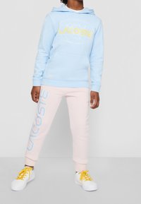 Ljusblå hoodie med gul "LACOSTE"-logga, mjukt tyg, känguruficka. Rosa mjukisbyxor med vit "LACOSTE"-text, elastiska muddar. Vita sneakers med gula snören.