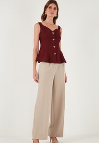 Burgundska peplum bluzica s V-izrezom, srebrnim dugmadima, uparena s bež širokim hlačama, postavljena na neutralnoj pozadini.