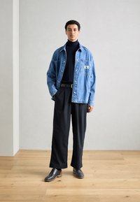 Jachetă din denim cu un buzunar pe piept, asortată cu pantaloni negri cu dungi subțiri. Un pulover negru cu guler înalt sub jachetă și pantofi gri completează ținuta.