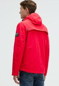 Homme aux cheveux châtain clair portant une veste à capuche rouge vif avec un patch de logo sur la manche et un jean bleu, tourné dos à la caméra.