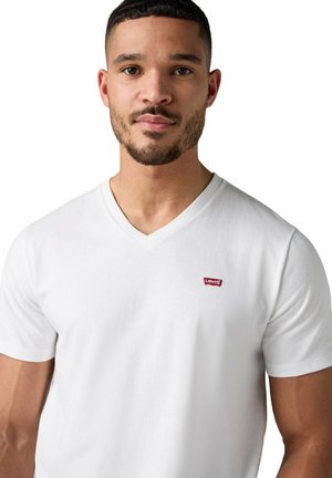 Mann trägt weißes Levi's T-Shirt mit V-Ausschnitt und rotem Logo-Patch auf der Brust, schaut nach vorne vor weißem Hintergrund.
