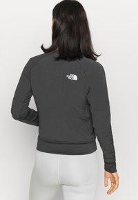 Vrouw met donker haar, gekleed in een zwarte lange mouw North Face-jack en witte broek, gezien van achteren tegen een effen achtergrond.