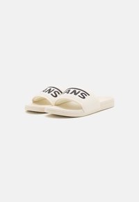 Sandale u boji krem s širokom trakom koja nosi crni "VANS" logo. Đon je ravan i napravljen od izdržljivog materijala.
