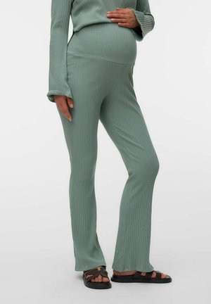 Kobieta w ciąży nosząca prążkowane spodnie ciążowe w kolorze szałwi zieleni i pasujący top, z czarnymi klapkami, stojąca na jednolitym tle.