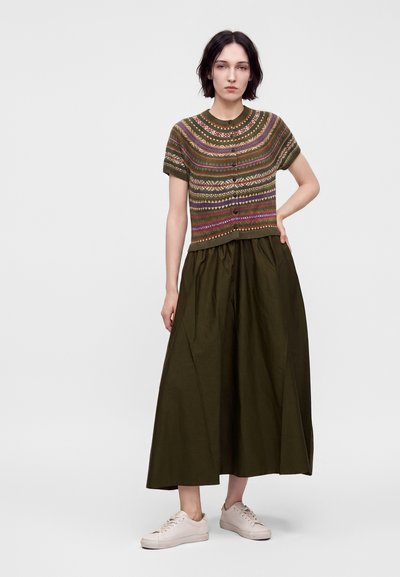 Femme aux cheveux courts et foncés portant un haut en maille à manches courtes à motifs, une longue jupe large sombre et des baskets claires, debout, main sur la hanche.